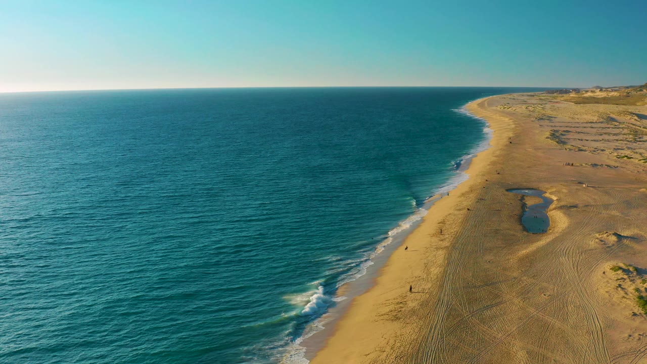 Aerial view over Diamante Cabo San Lucas beach where gentle surf reaches the sand and a small dark tide pool sits inland from the shoreline, with no visible people.