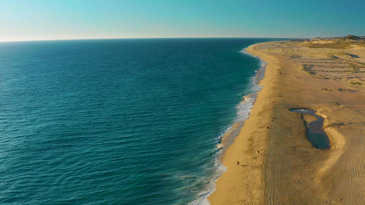 Drone view over Diamante Cabo San Lucas showing gentle white surf lines along the beach, textured sand on the right, open teal water on the left, distant dunes, and no visible people.