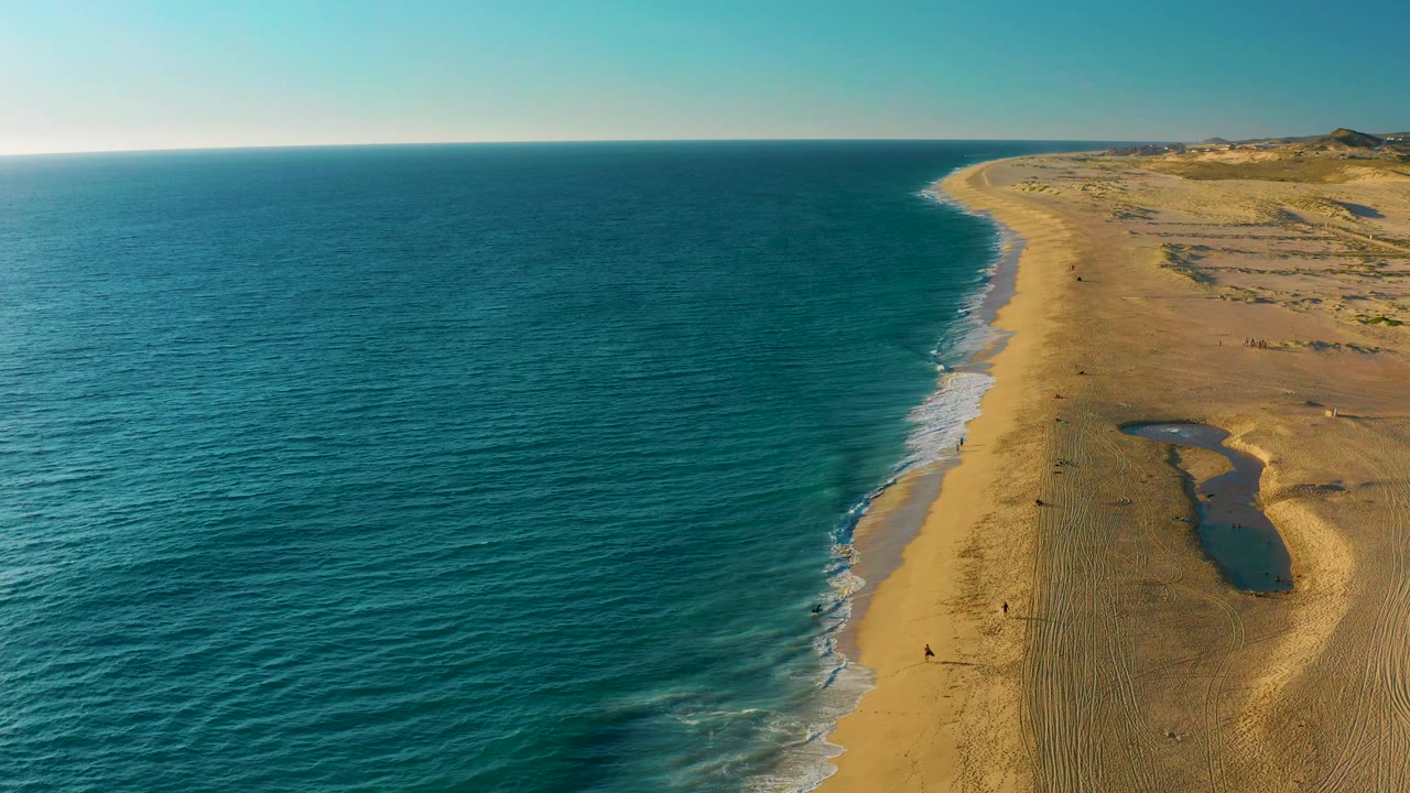 Aerial coastal frame at Diamante Cabo San Lucas with the shoreline bending toward the upper right, ocean texture filling the left side, a shallow dark pool on the sand, and no visible people.