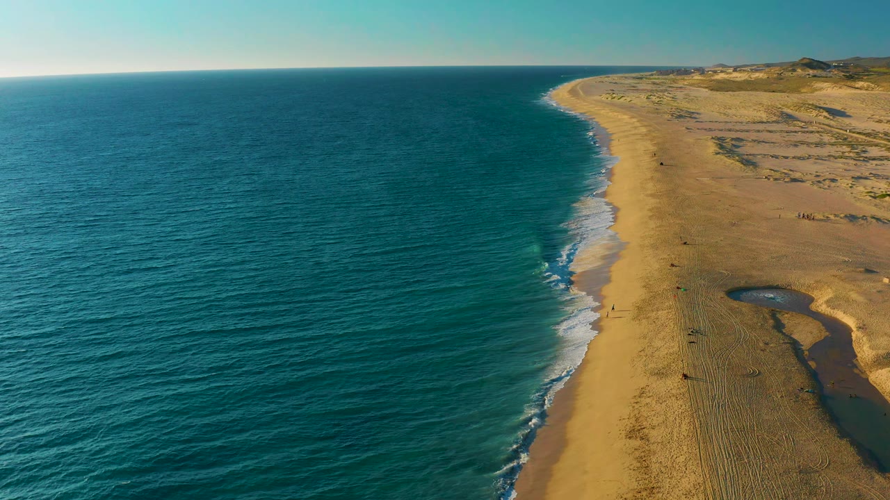 Aerial frame over Diamante Cabo San Lucas showing blue water pushing white foam closer to the golden beach, with dunes and sparse coastal structures in the distance and no visible people.