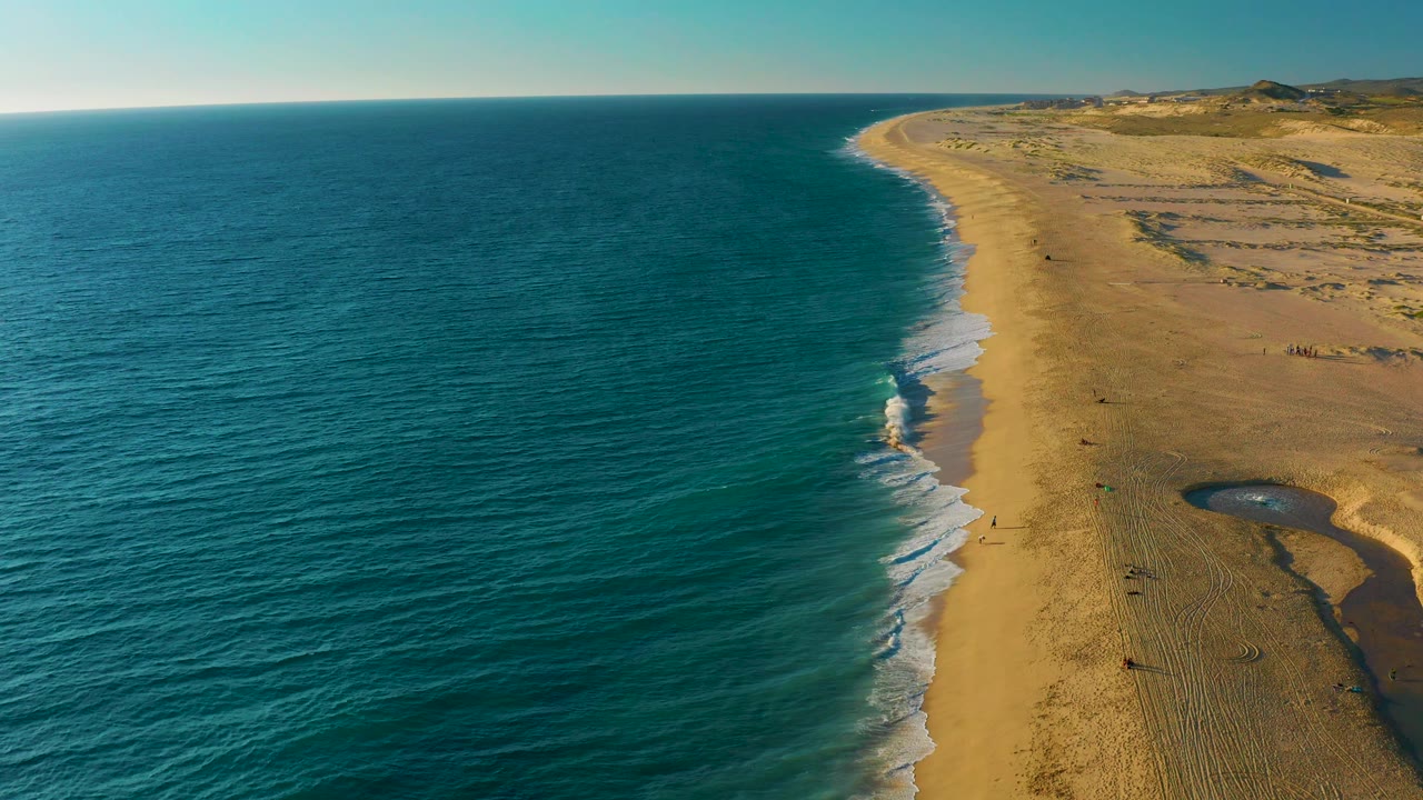 Aerial view above Diamante Cabo San Lucas with the blue ocean occupying most of the left frame, foam tracing the beach edge, broad sand and dunes on the right, and no visible people.