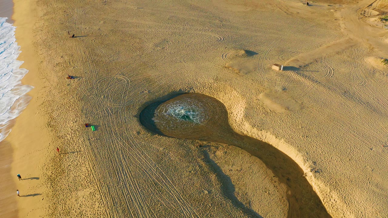 Aerial beach frame with dark sand pool, tracks, and surf line.