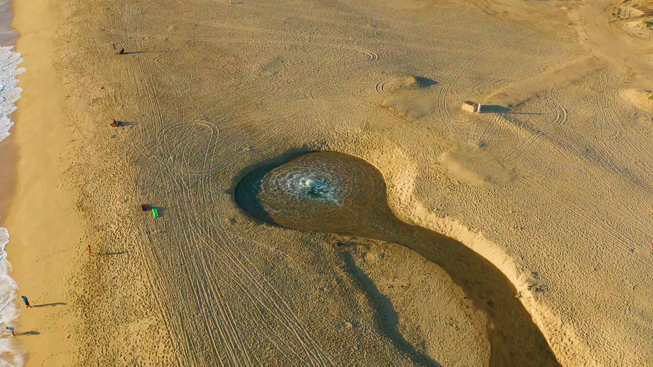 Overhead view at Diamante Cabo San Lucas showing a dark water-filled sand pool on golden beach sand, ocean foam along the left edge, visible tracks, and small distant people.