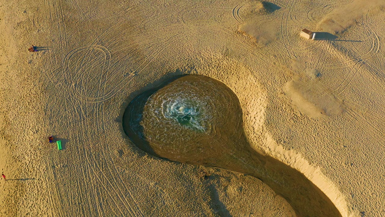 Overhead view in Diamante Cabo San Lucas showing a dark circular water feature in tan sand, tire tracks, small people at the left edge, and long shadows from objects on the beach.