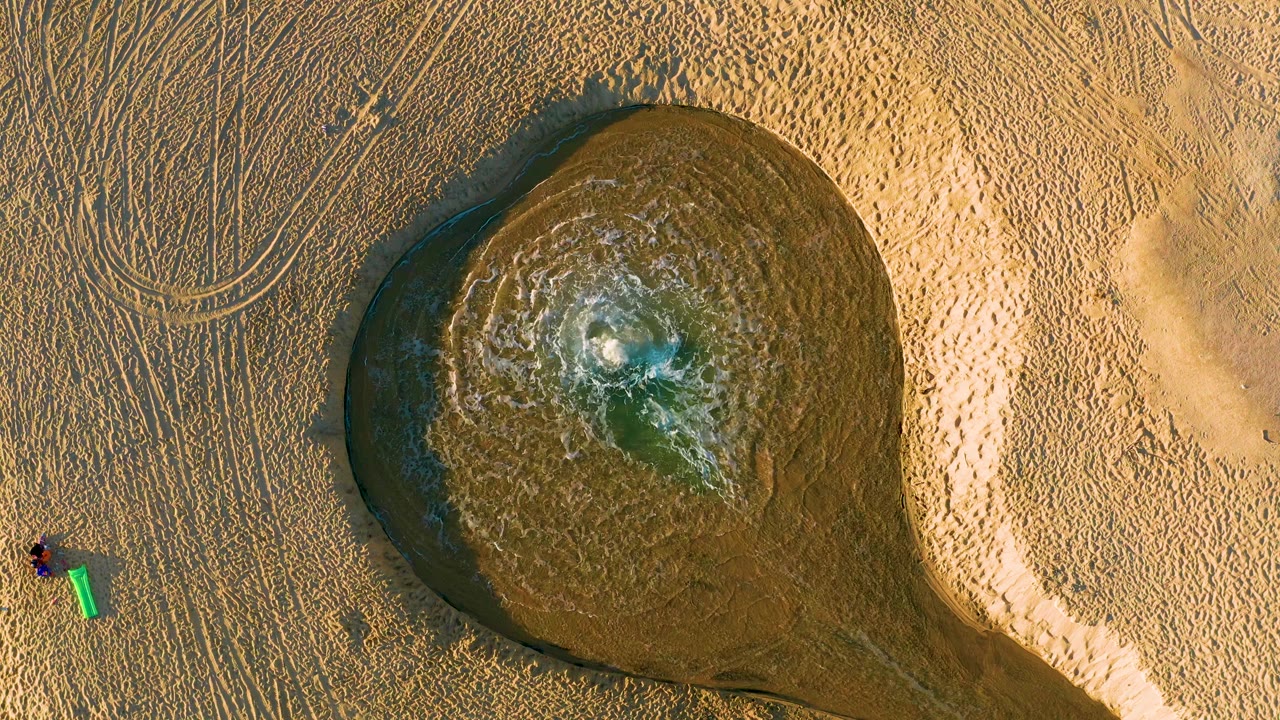 Muddy water pool with ripples, tire tracks, and a green bucket on sandy ground.