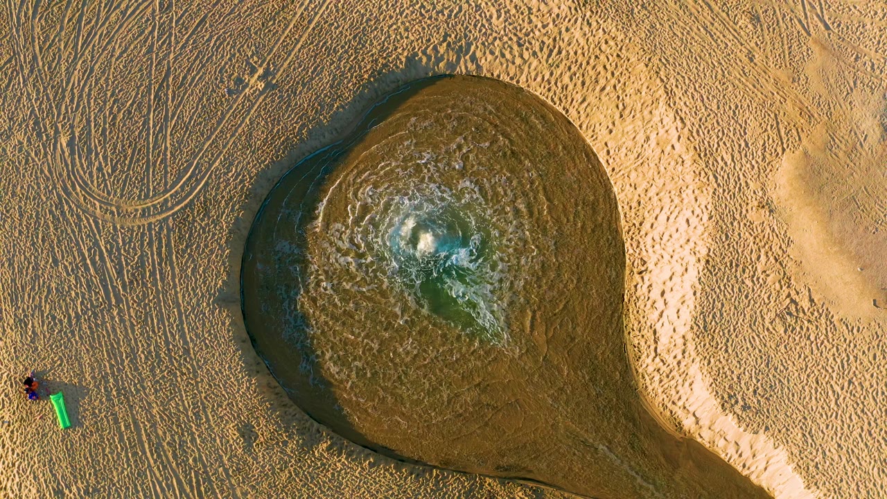 At 0:02, the muddy pool remains prominent from above, with disturbed water at its center and the person with the green bucket slightly separated on the surrounding sand.