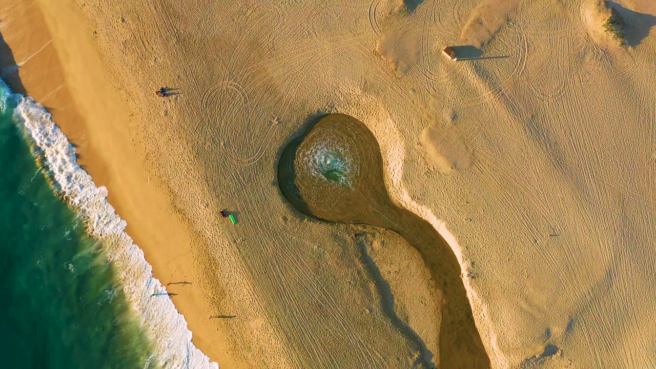 Overhead view of Diamante Cabo San Lucas beach with teal surf at the left edge, a rounded tide pool near center, raked tan sand, and tiny walkers near the shoreline.