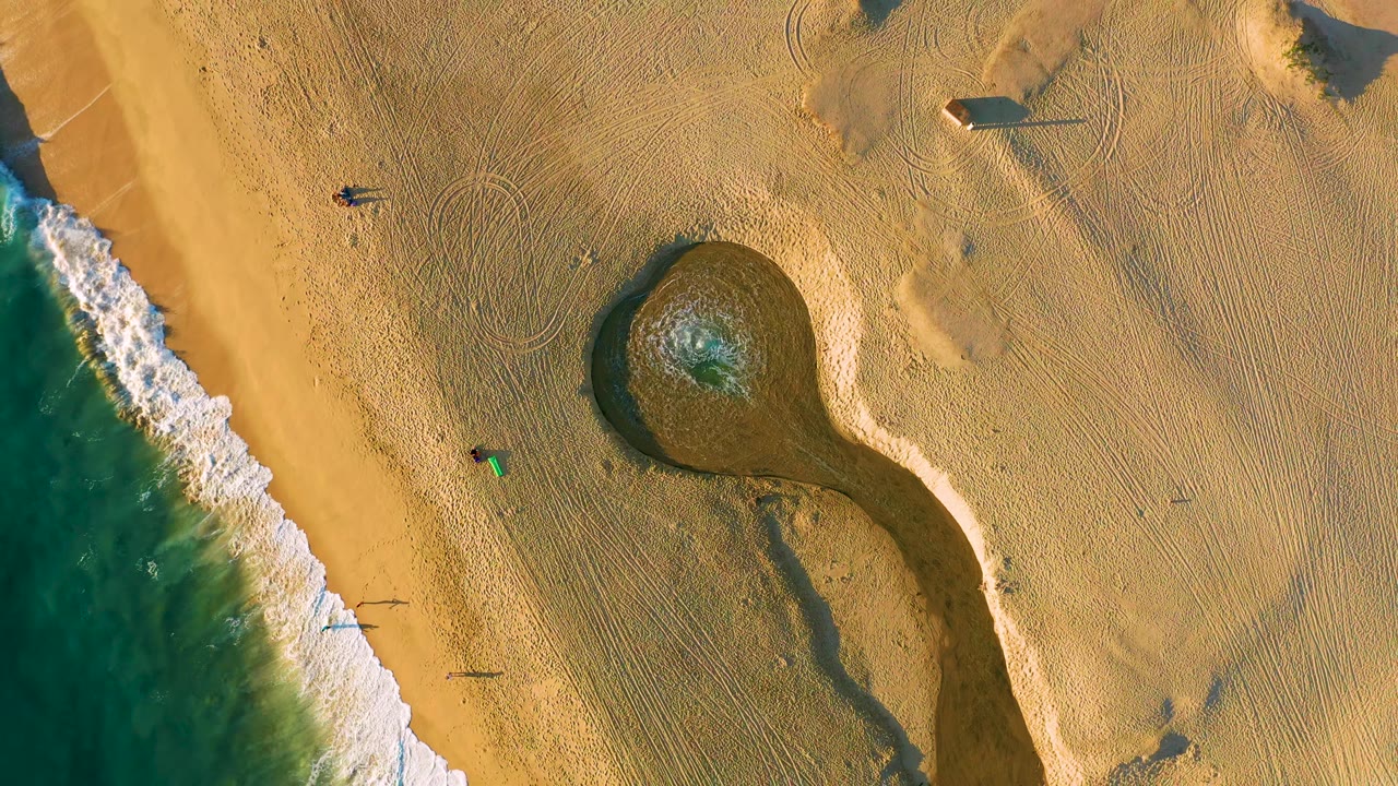Drone view of surf beside a rounded pool on raked sand.