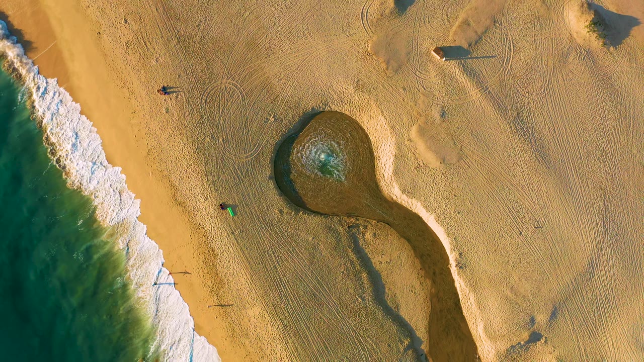 Aerial frame over Diamante Cabo San Lucas showing a broader band of white surf on the left, tan beach across the frame, and the dark rounded pool with rippled water near center.