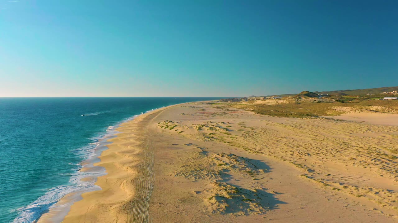 Aerial frame over Diamante Cabo San Lucas showing the shoreline curving toward distant hills, with small waves washing onto sand and no visible people.