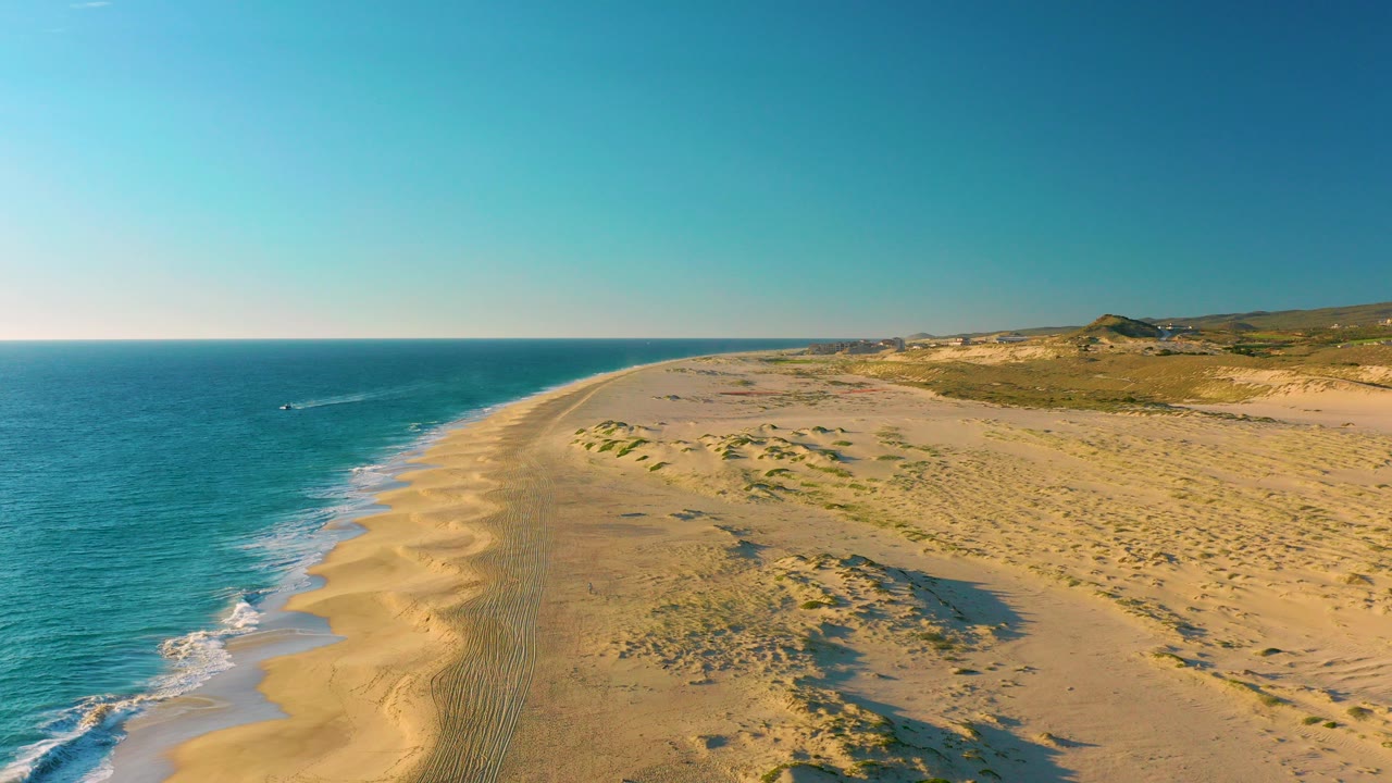 Aerial view over Diamante Cabo San Lucas showing blue ocean, a sandy shoreline, low dune ridges, distant hills, and no visible people at the start of the clip.