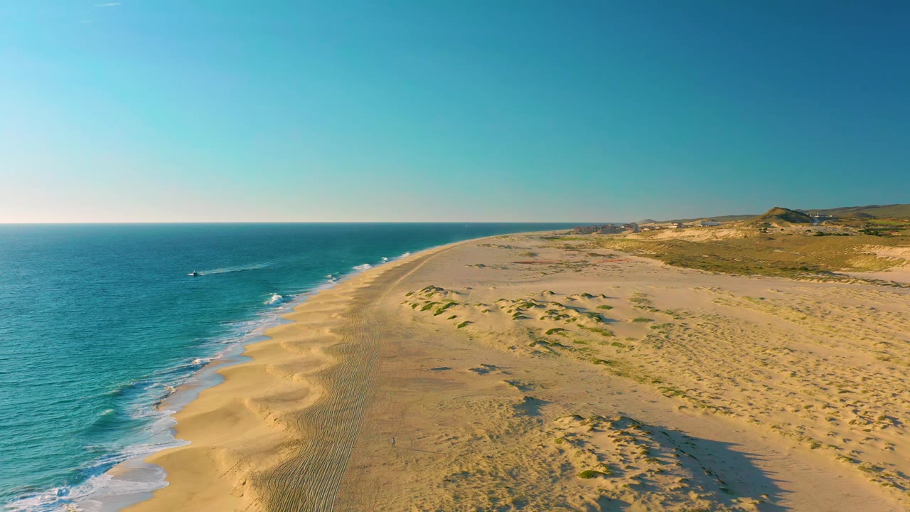 Drone view along the Diamante Cabo San Lucas shoreline, with the small boat farther out in blue water, low white waves at the beach, and no visible people.