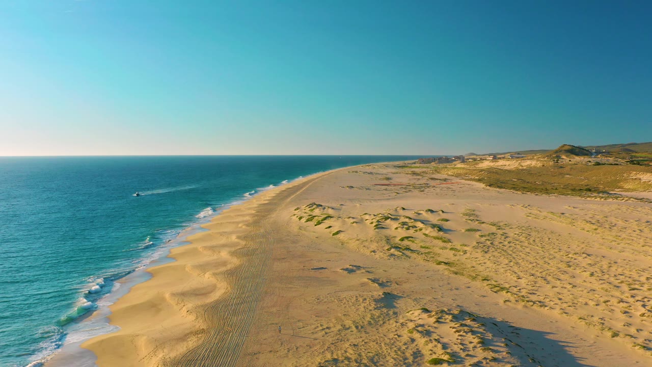 High drone view of the Baja California Sur coastline at Diamante Cabo San Lucas, with the boat closer to the left side of the frame, small waves on shore, and no visible people.