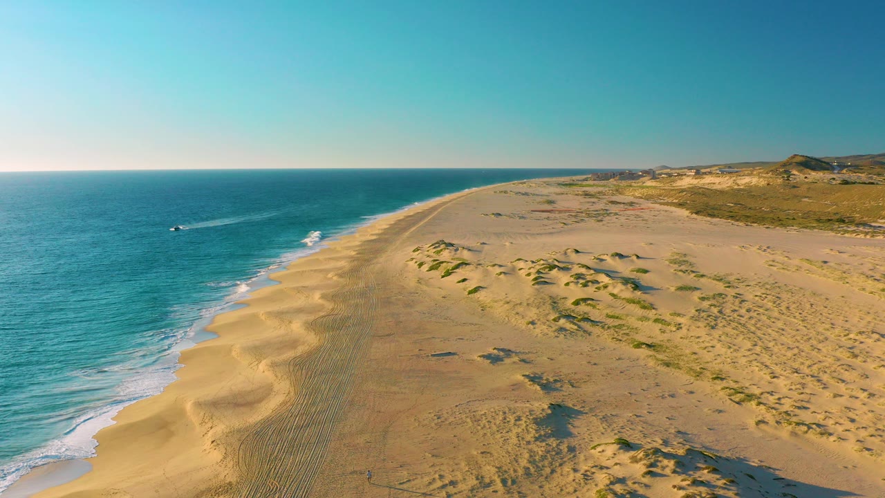 Aerial frame over Diamante Cabo San Lucas showing turquoise ocean, a long sandy beach, dune ridges, sparse vegetation, and a small boat offshore with no visible people.