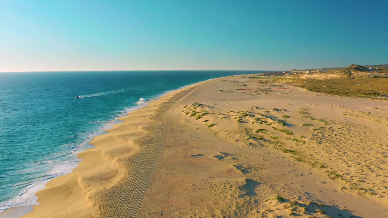 Elevated view of the Diamante Cabo San Lucas beach as the white boat wake stretches across the blue water, with pale sand, low dunes, inland scrub, and no visible people.