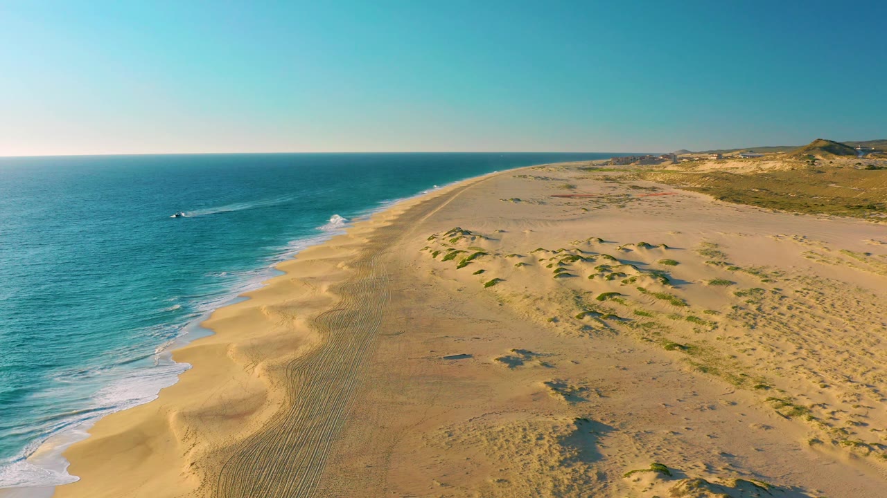 Aerial beach and ocean view with small boat offshore and dune field inland.