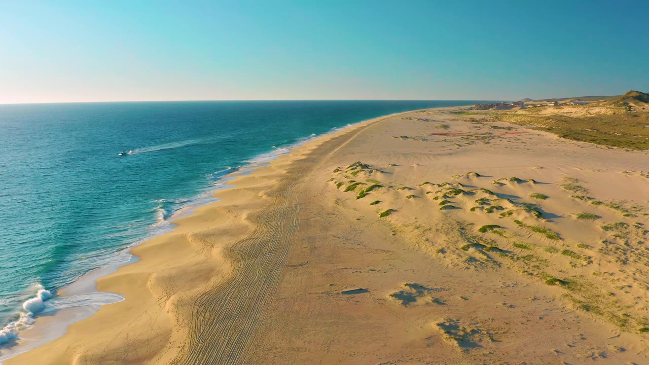 Drone view along Diamante Cabo San Lucas shoreline as the speedboat wake stretches farther across the blue water beside broad sand dunes, with no visible people.