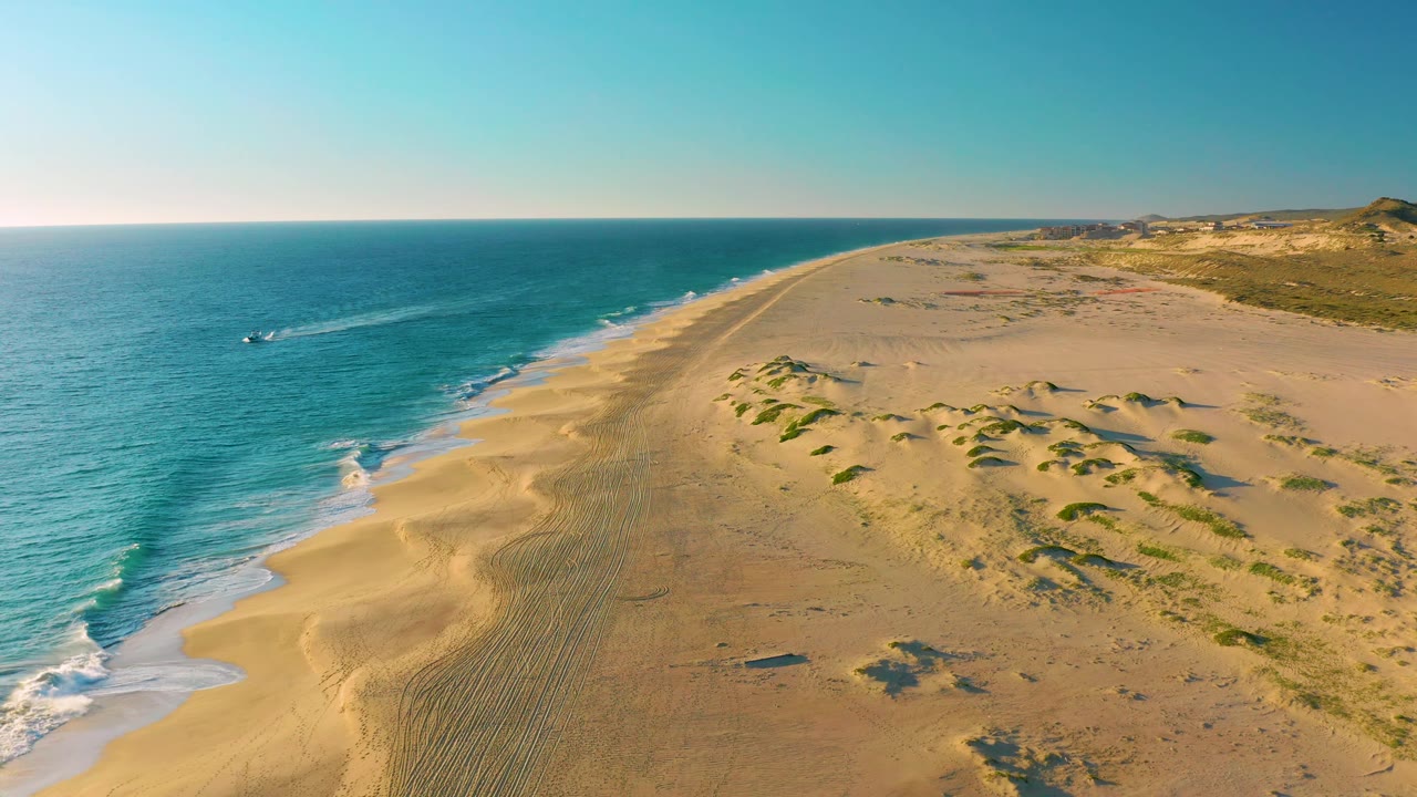 Wide aerial frame of Diamante Cabo San Lucas coastline with the boat and its wake slightly farther along the left-side ocean while dunes and beach tracks remain visible, with no visible people.
