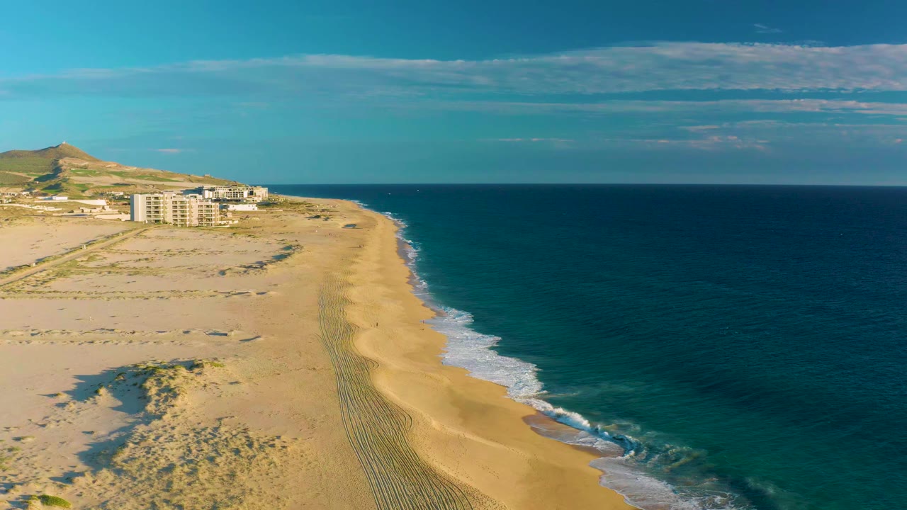 Opening aerial frame over Diamante Cabo San Lucas beach, with dunes and tire tracks in the sand, low buildings near a hill, blue ocean to the right, and no visible people.