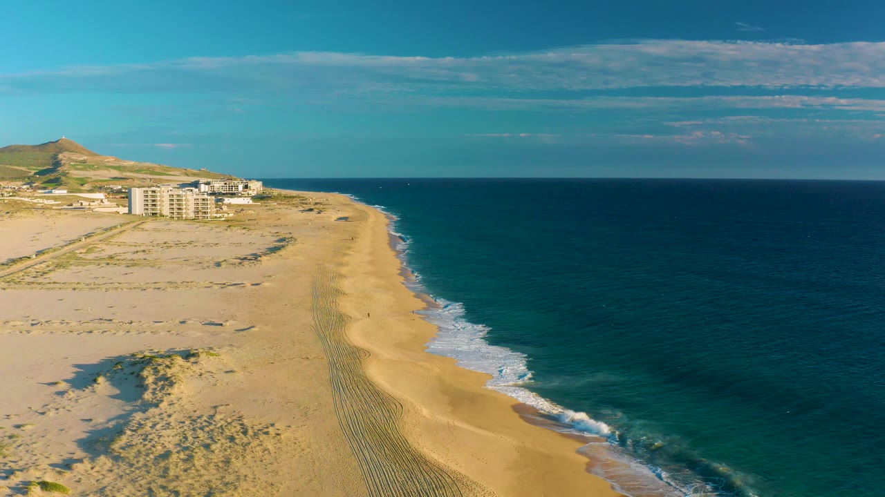 Drone perspective along the Diamante Cabo San Lucas coast, with low buildings left of center, the sandy shoreline curving away, white wave foam on the beach, and no visible people.