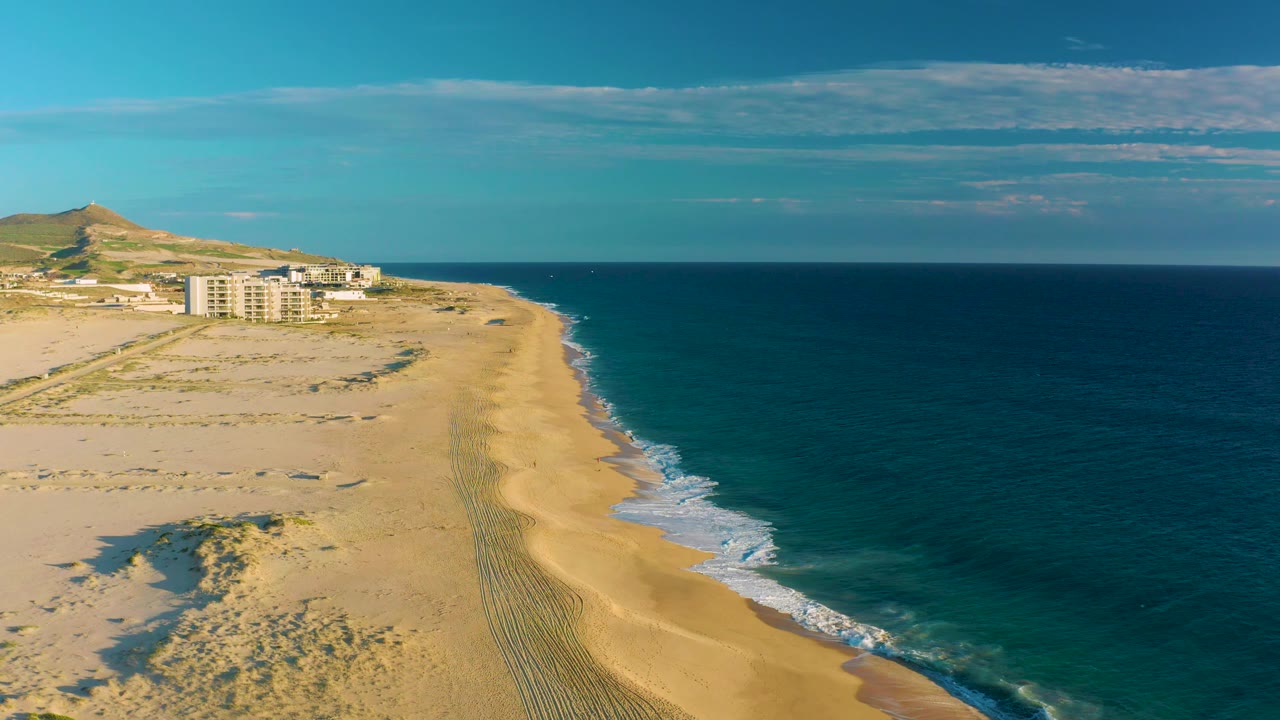 Aerial shoreline with dunes, tire tracks, distant buildings, and calm surf.