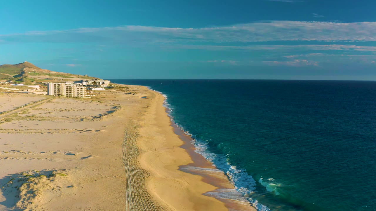 At 0:01, the Diamante Cabo San Lucas shoreline remains centered, with surf edging the sand, ocean filling the right side, buildings on the left, a low hill behind them, and no visible people.