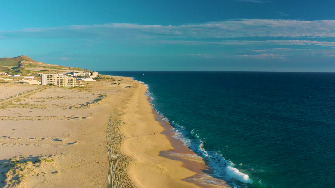 At 0:02, the aerial view shows a slightly tighter read of the curving sandy shoreline at Diamante Cabo San Lucas, with white waves, open ocean, distant buildings, hill, and no visible people.