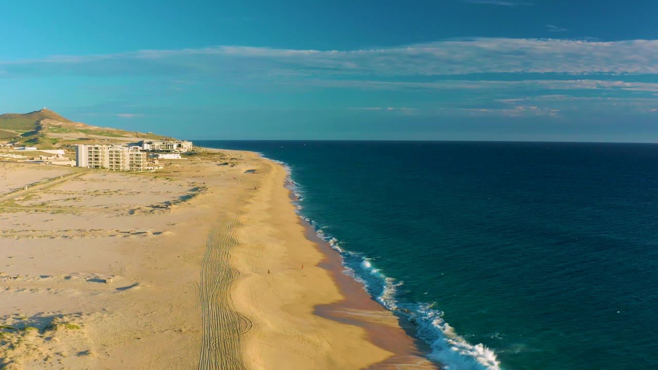 At 0:03, the high coastal view shows more foreground dunes and sand tracks, the surf running diagonally beside the beach, distant buildings, the hill, and no visible people.