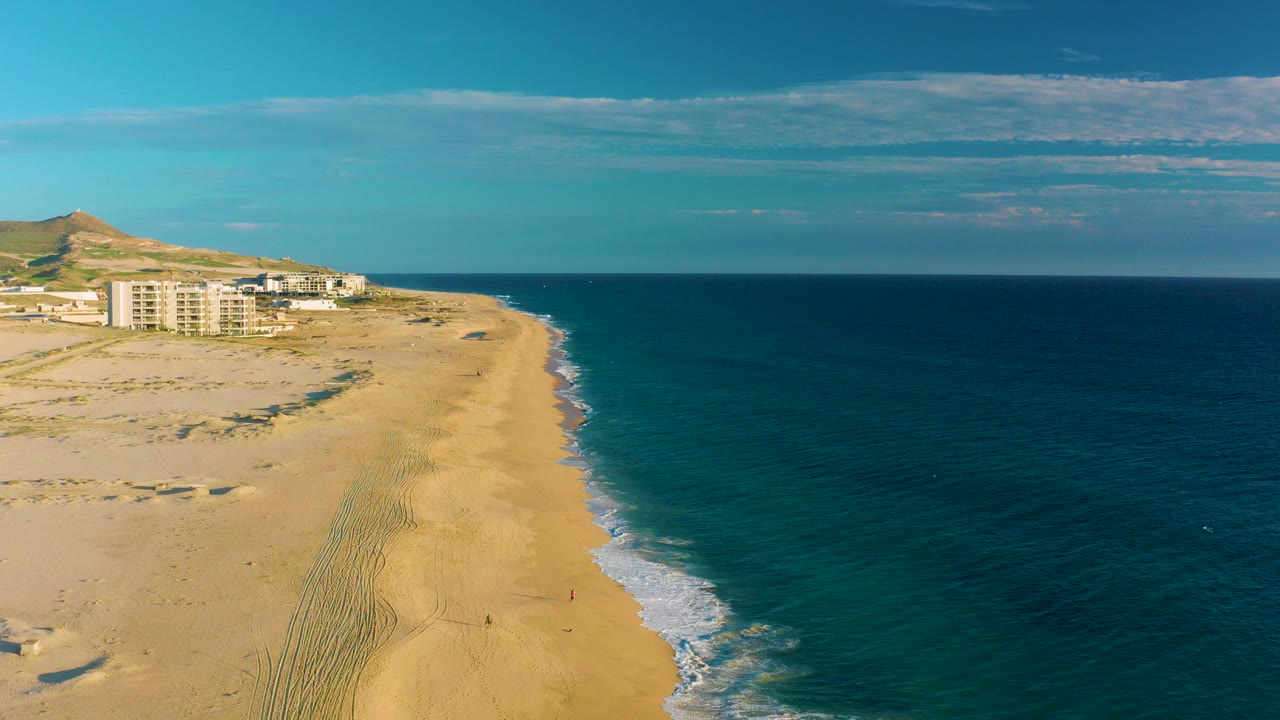 Opening aerial frame over Diamante Cabo San Lucas shows a wide sandy beach beside blue ocean, distant coastal buildings, a low hill, thin clouds, and no visible people.