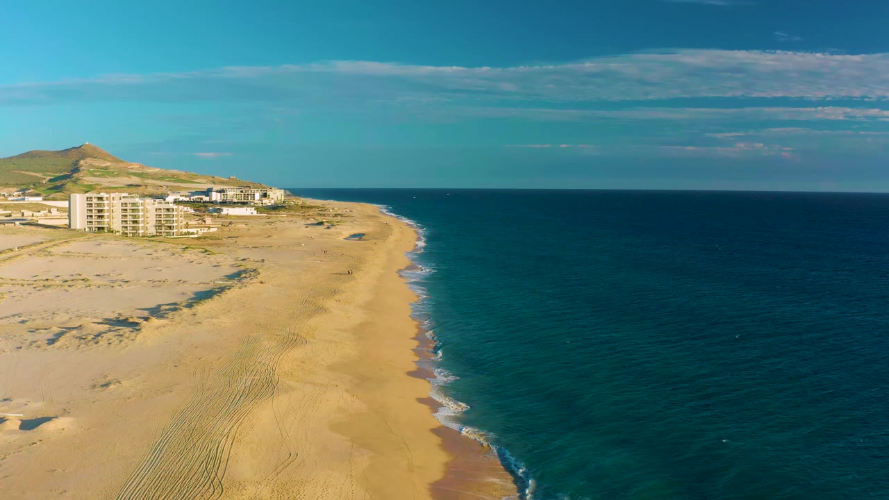 Sandy coastline with turquoise water, waves, buildings, and hill in the distance.
