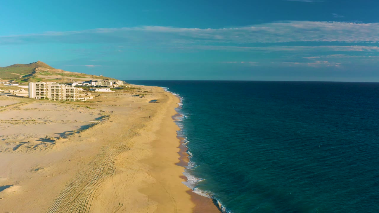 Drone perspective over Diamante Cabo San Lucas with empty sand filling the foreground, ocean along the right edge, pale buildings below the hill, and no visible people.
