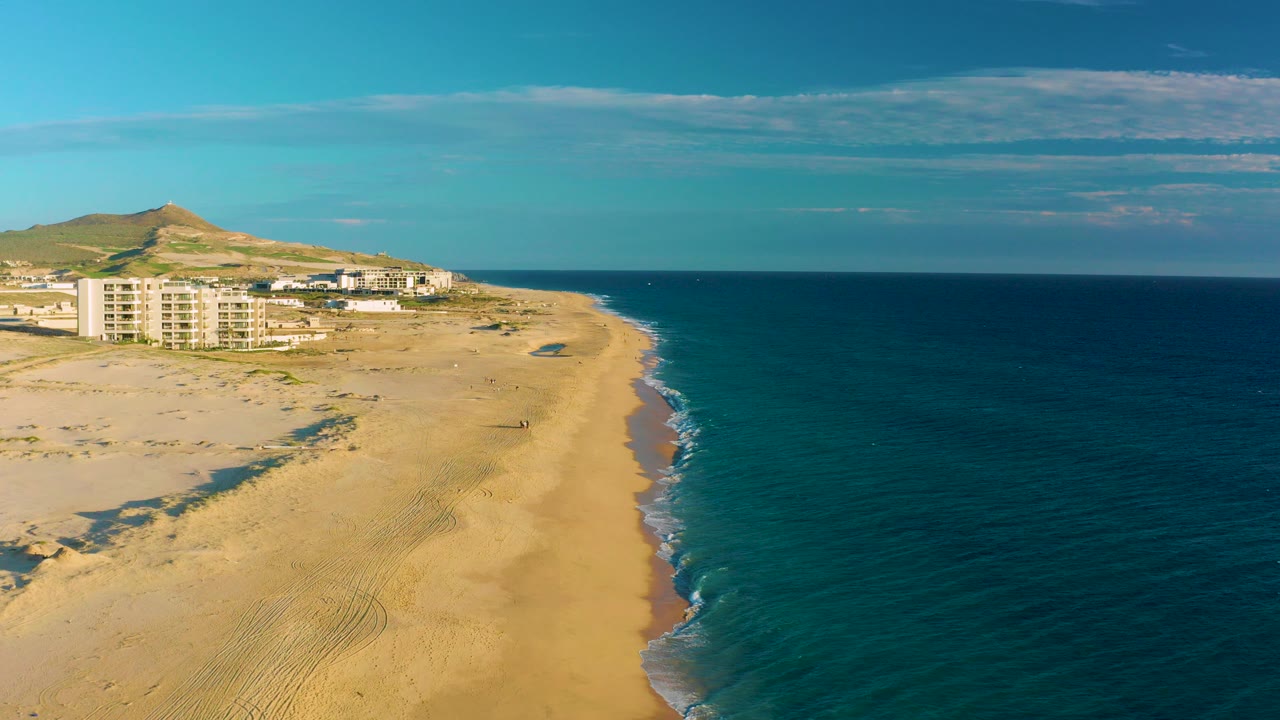 Aerial frame over Diamante Cabo San Lucas showing a sandy beach curving beside dark blue ocean, pale coastal buildings, dry hills, clear sky, and no visible people at the opening moment.