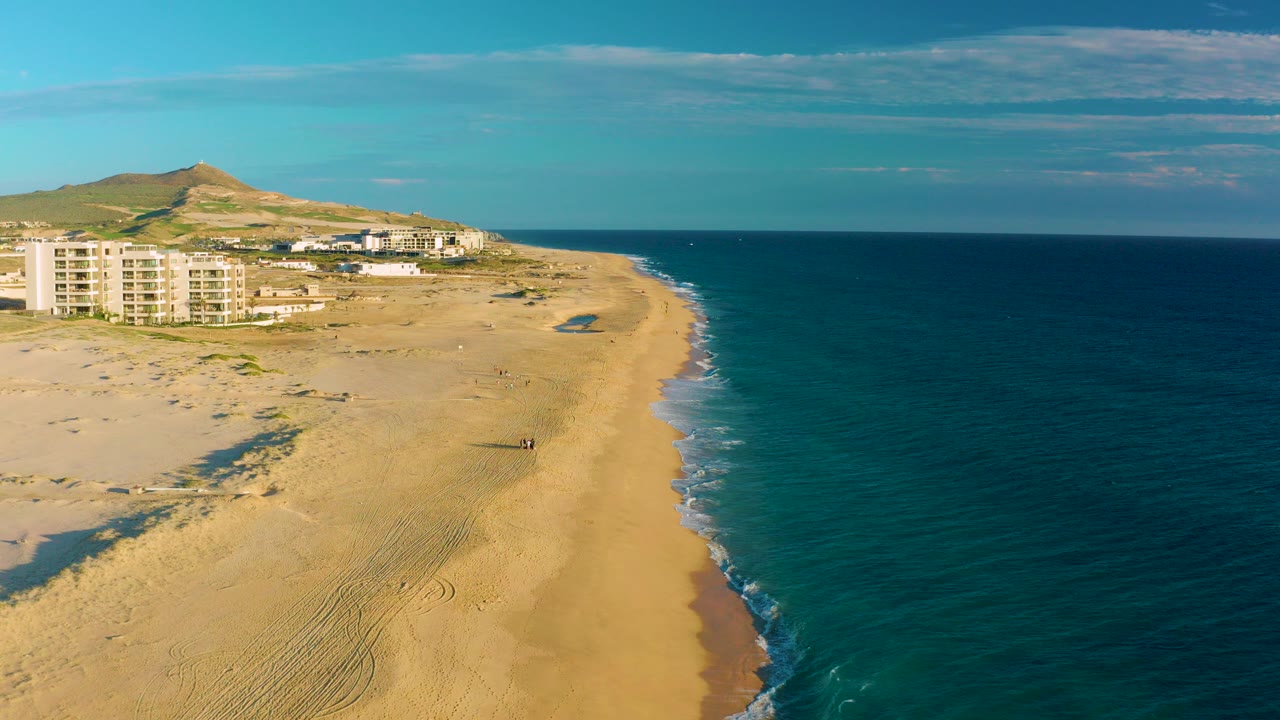 Aerial view over Diamante Cabo San Lucas with wide sand at left, blue ocean at right, beachside buildings near the horizon, a hill behind them, and a small person visible on the beach.