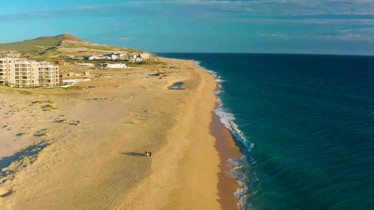Drone view of beach and surf with walkers casting shadows near coastal buildings.