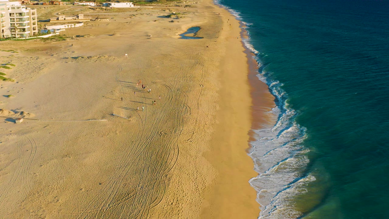 High aerial view over Diamante Cabo San Lucas with a wide sandy beach, blue ocean along the right edge, faint beach tracks, small people near the center, and buildings at the upper left.