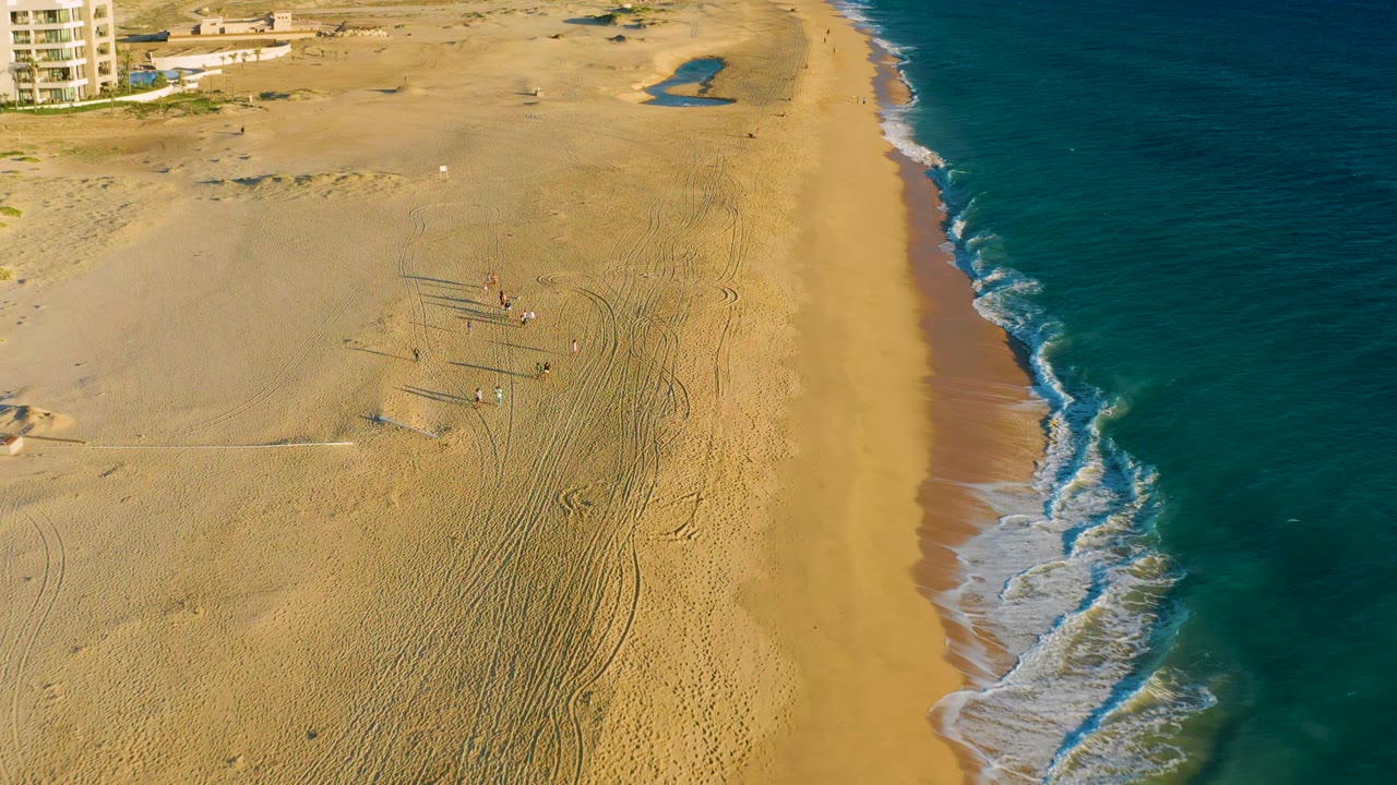 Overhead view of Diamante Cabo San Lucas beach with the ocean occupying the right side, people grouped around objects on the sand, vehicle-like tracks, and warm light across the beach.