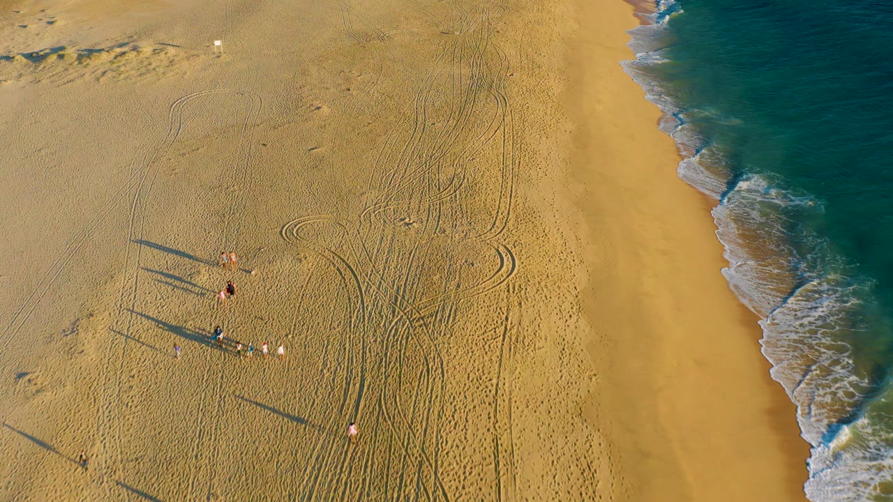 Aerial view over Diamante Cabo San Lucas beach with a small group of people on golden sand, long shadows, tire tracks, and blue surf along the right edge.