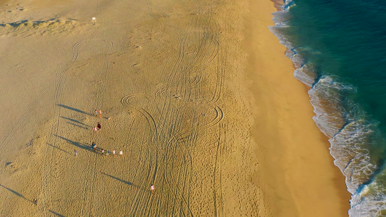 Small people on a sandy beach near tire tracks and blue ocean water.
