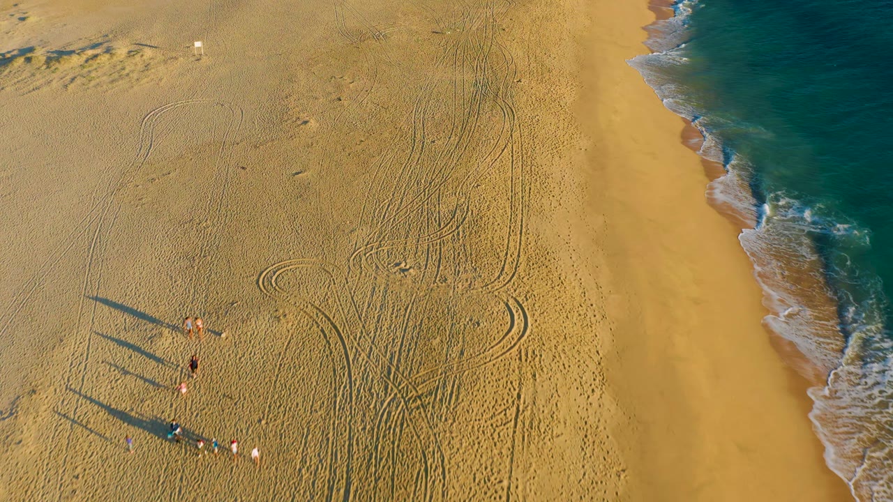Aerial view of Diamante Cabo San Lucas sand with the group shifted lower in frame, dark elongated shadows, tire marks, and a strip of turquoise ocean surf to the right.