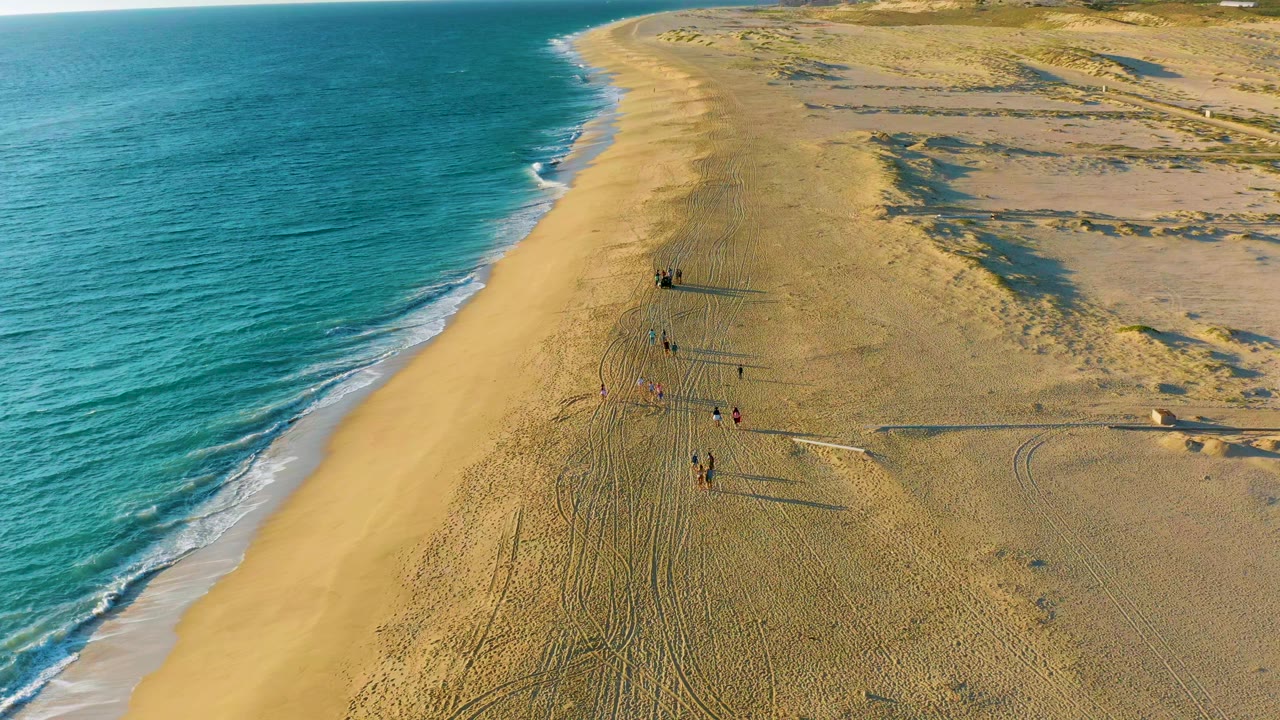 Aerial frame over Diamante Cabo San Lucas showing turquoise water on the left, a broad sandy shore, tire tracks, dunes, and small visible people walking near the center.