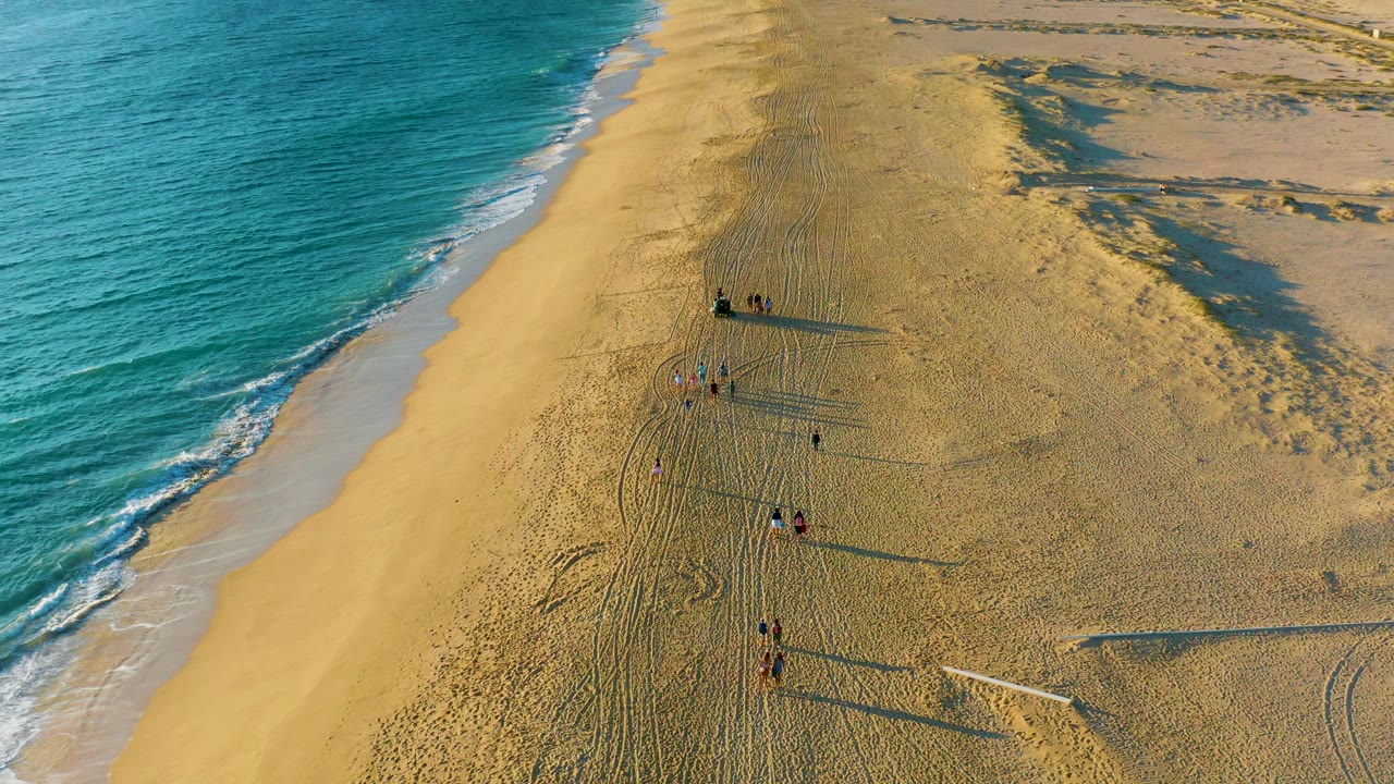 Aerial frame over Diamante Cabo San Lucas showing blue ocean on the left, broad sand on the right, and several distant people walking with long shadows.