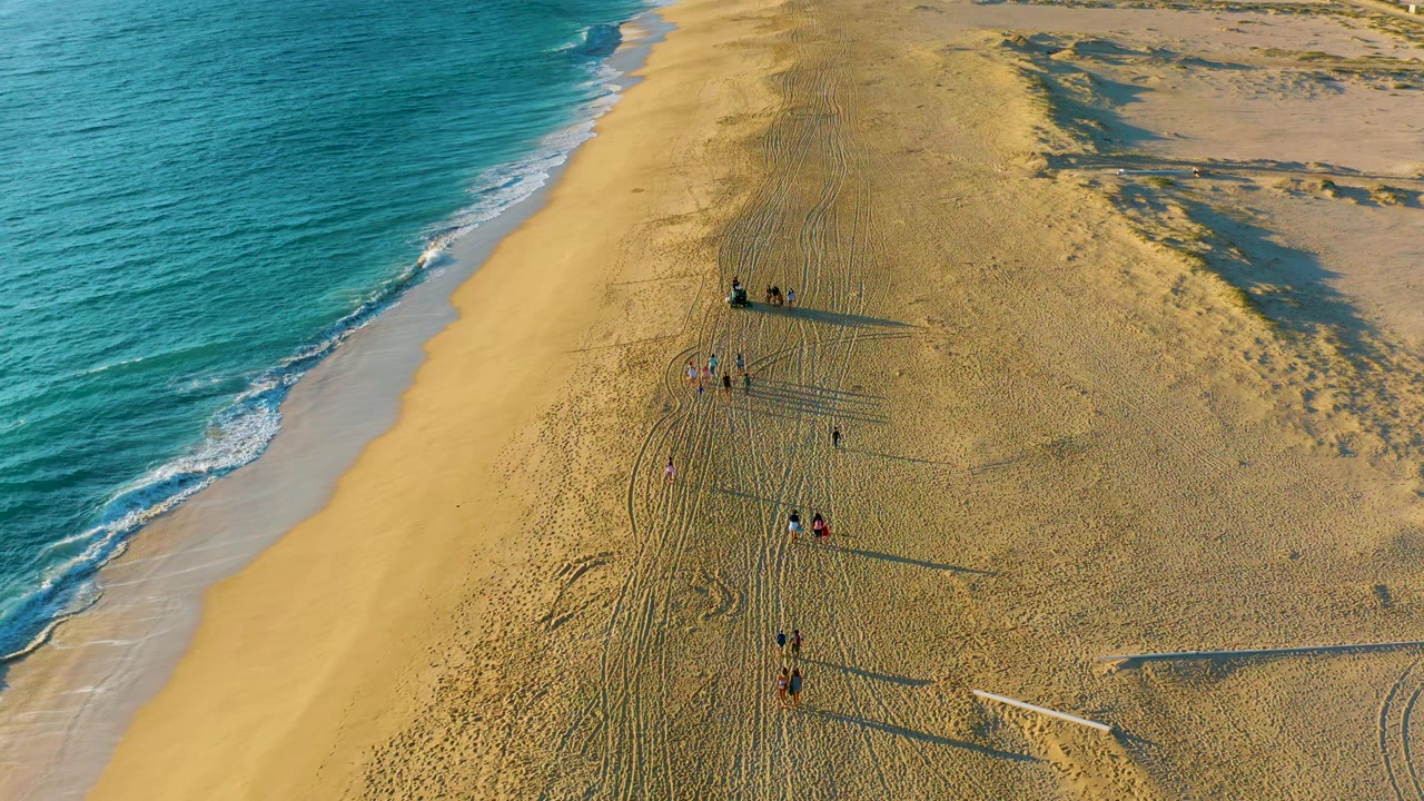 High aerial frame over the Diamante Cabo San Lucas beach showing teal water, pale surf, tire tracks in the sand, dunes, and distant visible people.