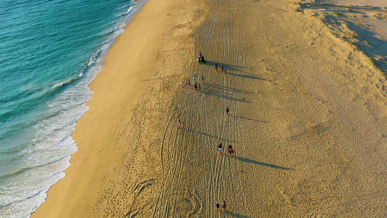 Drone-height frame of Diamante Cabo San Lucas beach where the wave line sits left, several people stand or walk near tracks, and their shadows extend across the sand.