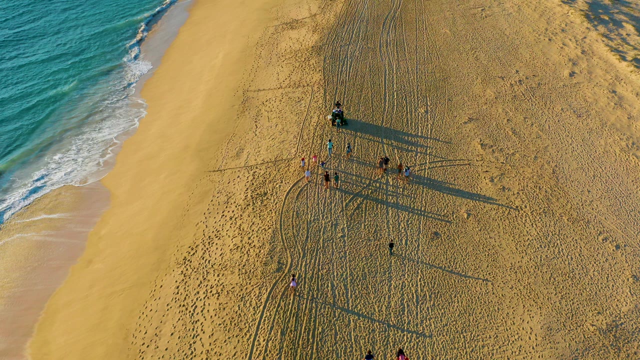 Overhead drone frame of Diamante Cabo San Lucas beach with turquoise surf at left, yellow sand, tire tracks, footprints, and a small group of people casting long shadows near the center.