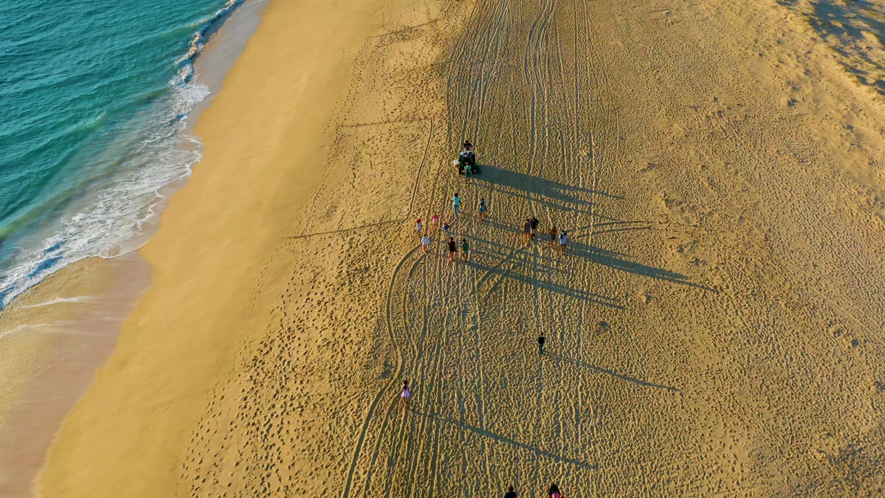 Aerial view of walkers on sandy beach beside blue ocean.