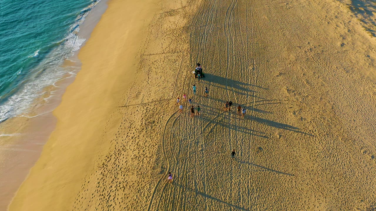 High overhead frame of the Diamante Cabo San Lucas shore where several people move across marked sand, with foamy waves entering from the upper left and shadows extending to the right.