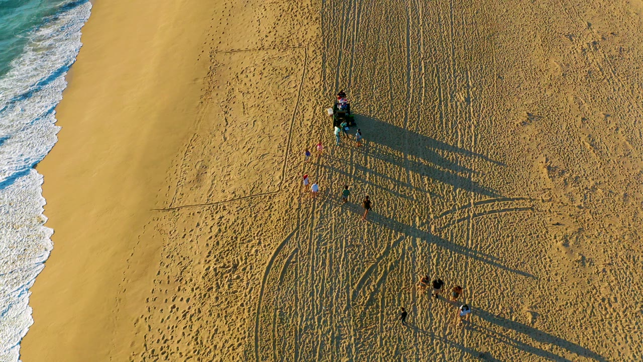 Overhead view of a Diamante Cabo San Lucas beach with ocean foam on the left, a small vehicle and people on rippled sand, and long shadows stretching to the right.