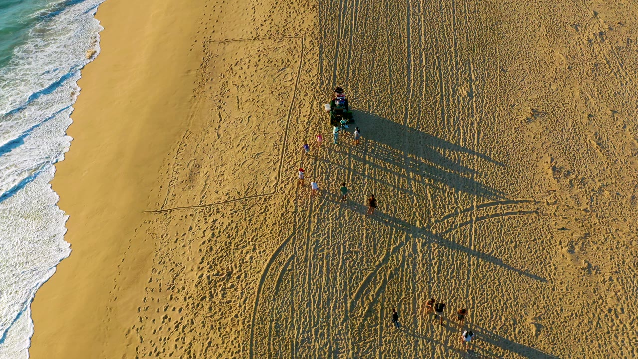 Drone-style overhead view at Diamante Cabo San Lucas with a brighter band of surf on the left, the vehicle and people farther right, and long shadows crossing textured sand.
