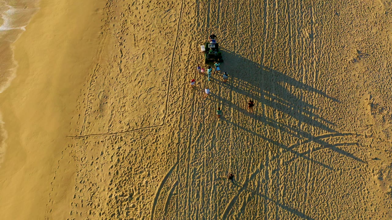 Drone-style overhead view of beachgoers near a vehicle on tracked sand by the ocean.