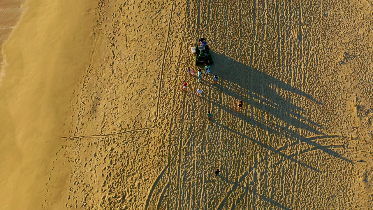 At 0:02, the vehicle and visible people sit near the upper right portion of the sandy beach, while curved tire marks and long shadows cross the Diamante Cabo San Lucas shoreline scene.